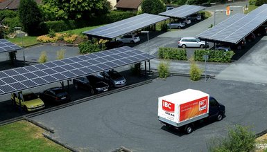 Schotterparkplatz mit Carports, die Solarmodule auf dem Dach haben; im Vordergrund ein kleiner Laster, der mit IBC SOLAR Werbung bedruckt ist