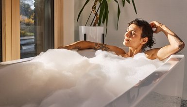 Entpannt aussehende Frau mit Tattoos auf den Armen in einer Badewanne voller Schaum, im Hintergrund ein Fenster und eine Zimmerpflanze