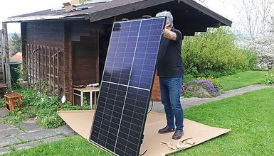 Mann steht vor einem Gartenhaus und hält ein Solarmodul auf dem Rasen