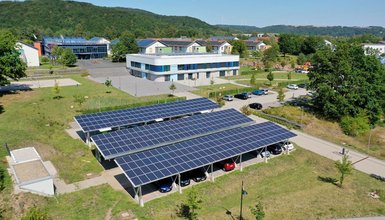 Solar-Carports mit Photovoltaikanlagen auf Parkplätzen vor einem Bürogebäude; im Hintergrund Wohnhäuser mit Solardächern.