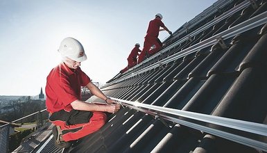 Zwei Handwerker in roter Kleidung und weißen Helmen die auf einem Dach im Sonnenschein eine Solaranlage montieren