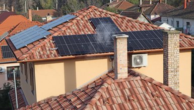 Mehrere Hausdächer aus der Vogelperspektive, auf zwei Dächern befinden sich Solarmodule, ein Schornstein im Vordergrund raucht und an der Wand eines Hauses ist eine Klimaanlage montiert