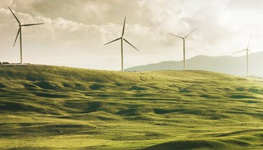 Grüne hügelige Landschaft, auf Erhebung stehen vier Windräder, die sich zu drehen scheinen
