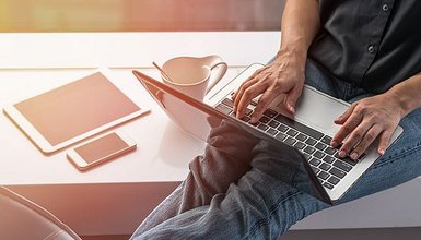 Nahaufnahme einer Person am Laptop, daneben liegen ein Tablet, ein Handy und eine Tasse.