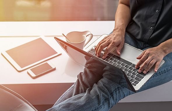 Nahaufnahme einer Person am Laptop, daneben liegen ein Tablet, ein Handy und eine Tasse.